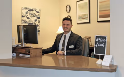 A man in a suit sitting behind a reception desk in an office area. The desk has a computer monitor, a small sign that says 'Welcome friends', and some business cards. The background features a lamp, a clock, and framed artwork on the wall.