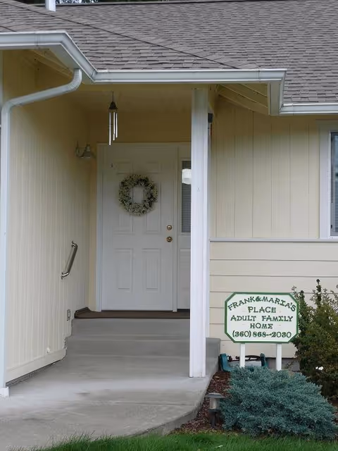 Entrance to a beige adult family home with a white door decorated with a green wreath. There is a small porch with a handrail on the left side and a sign on the right that reads 'Frank & Maria's Place Adult Family Home (360) 869-2030'.