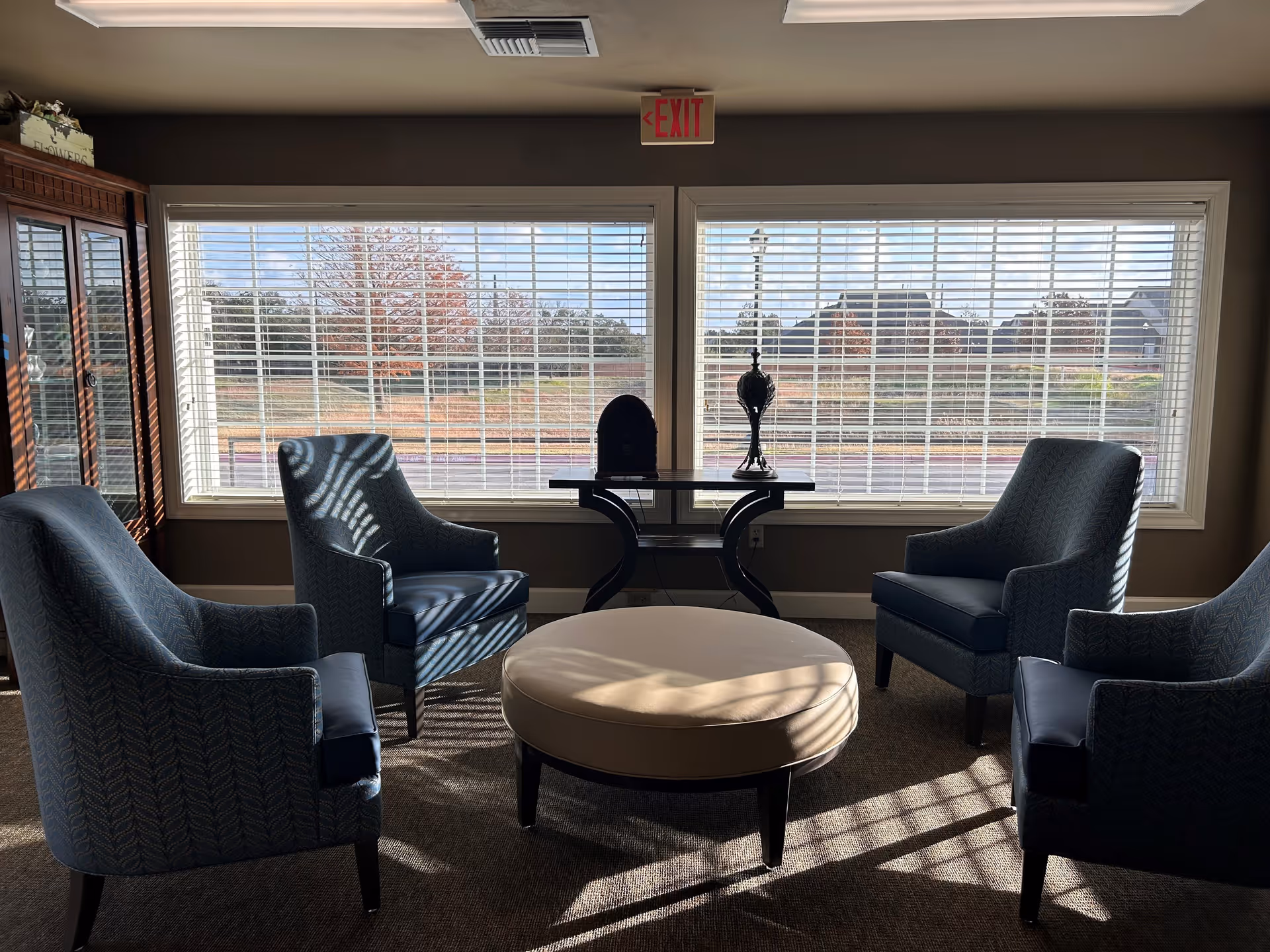 A cozy sitting area with four blue upholstered armchairs arranged around a round beige ottoman. Behind the seating is a large window with white blinds partially open, letting in natural light and showing an outdoor view with trees and houses. A small table with decorative items is placed between the windows, and an exit sign is visible above the window.