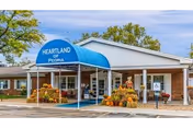 Front entrance of a senior living facility with a blue awning reading "HEARTLAND OF PEORIA" and fall pumpkin decorations.