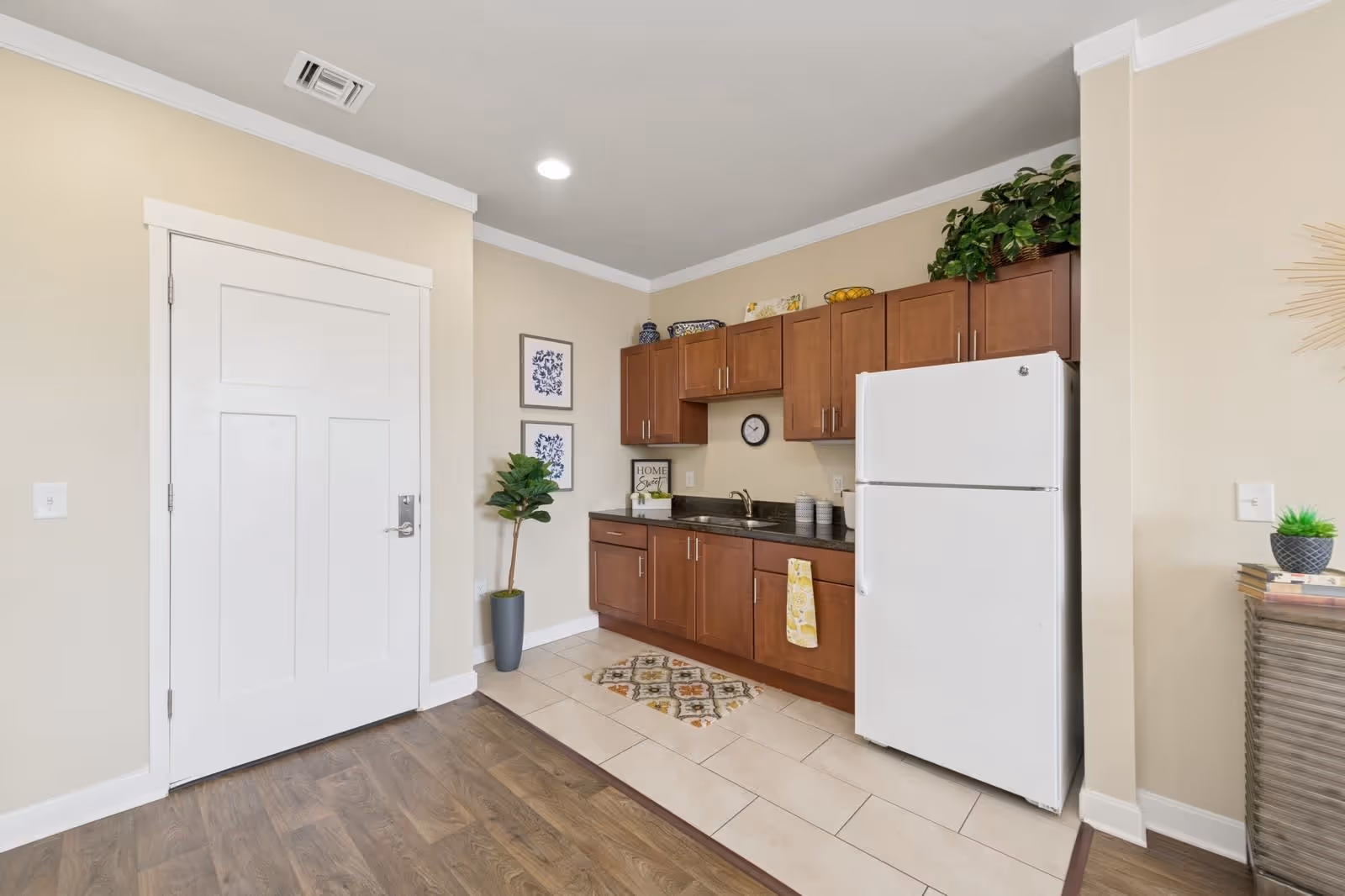 A small kitchen area with wooden cabinets, a white refrigerator, a black countertop with a sink, and a yellow towel hanging from a cabinet handle. The floor is a combination of wood and tile. There is a white door on the left side, two framed pictures on the wall, a potted plant, and decorative items on top of the cabinets.