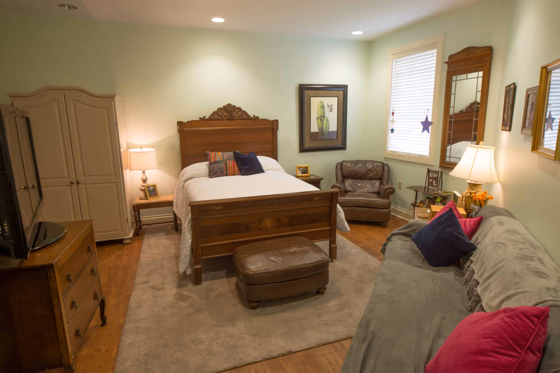 Cozy furnished bedroom with a wooden bed, armchair, sofa, dresser, and TV.