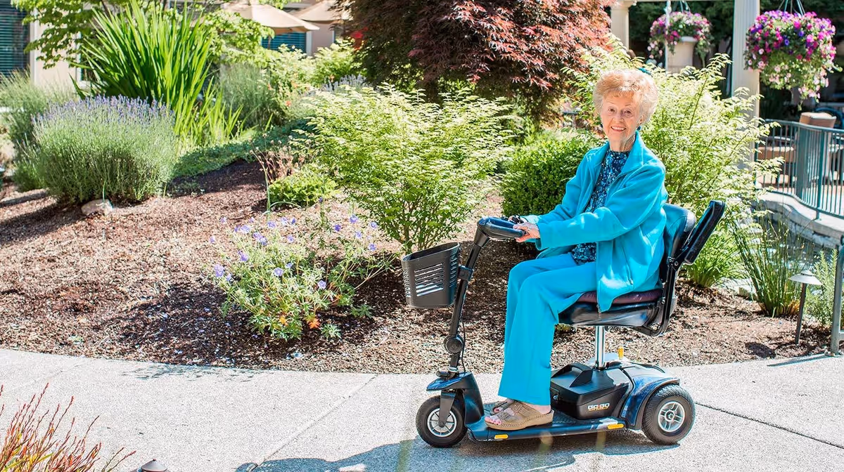An elderly woman wearing a turquoise outfit is sitting on a mobility scooter on a paved path in a garden area with various green plants and flowers around her. She is smiling and appears to be enjoying the outdoor setting.