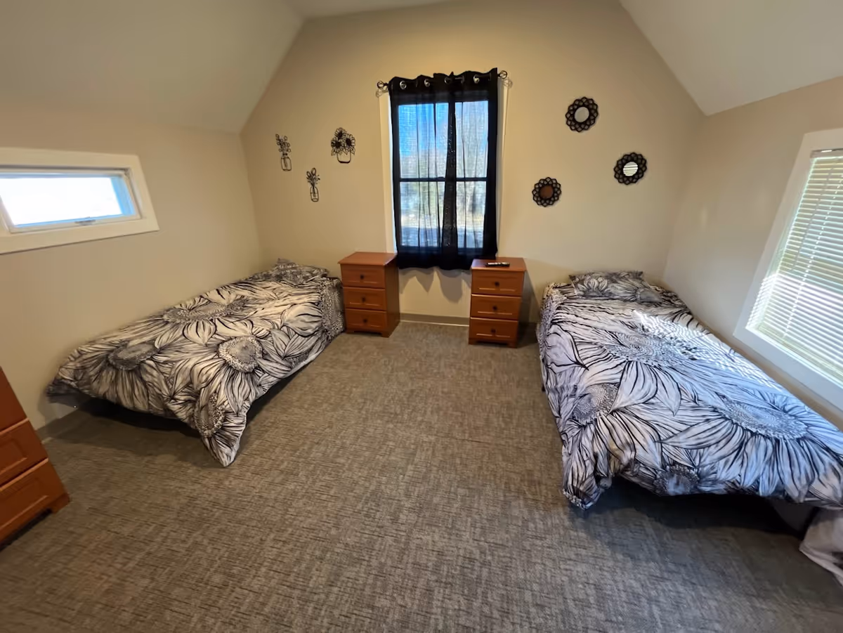 A bedroom with two single beds placed against opposite walls, each with black and white floral patterned bedding. Between the beds are two wooden nightstands with drawers. A window with black curtains is centered on the wall behind the nightstands. The walls are decorated with small black metal wall art and round mirrors. The room has carpeted flooring and neutral-colored walls.