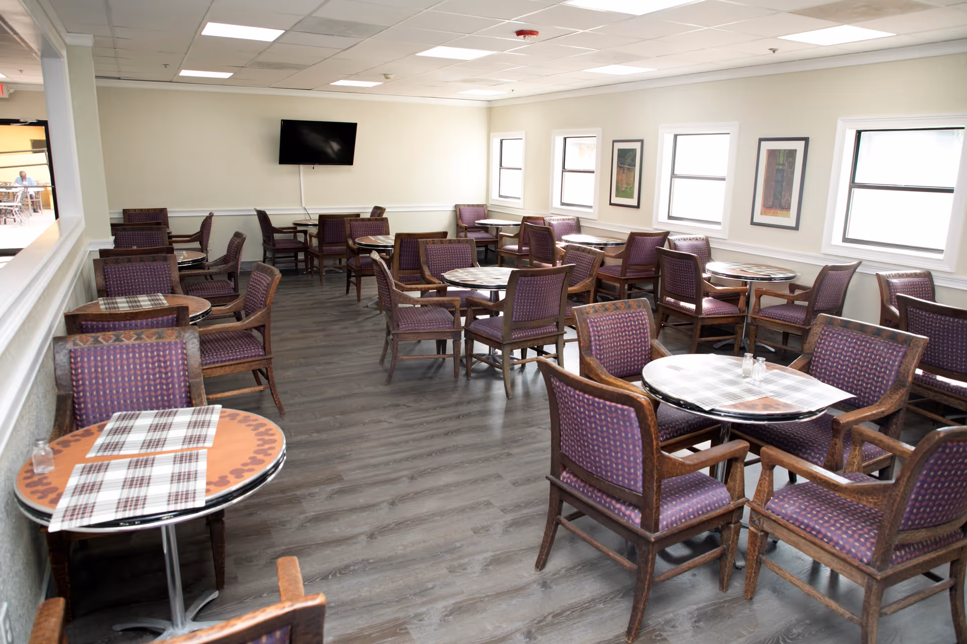 A bright dining room with multiple round tables covered with checkered placemats and surrounded by wooden chairs with purple patterned cushions. The room has light-colored walls, several windows letting in natural light, framed artwork on the walls, and a flat-screen TV mounted on one wall. The floor is a dark wood laminate.