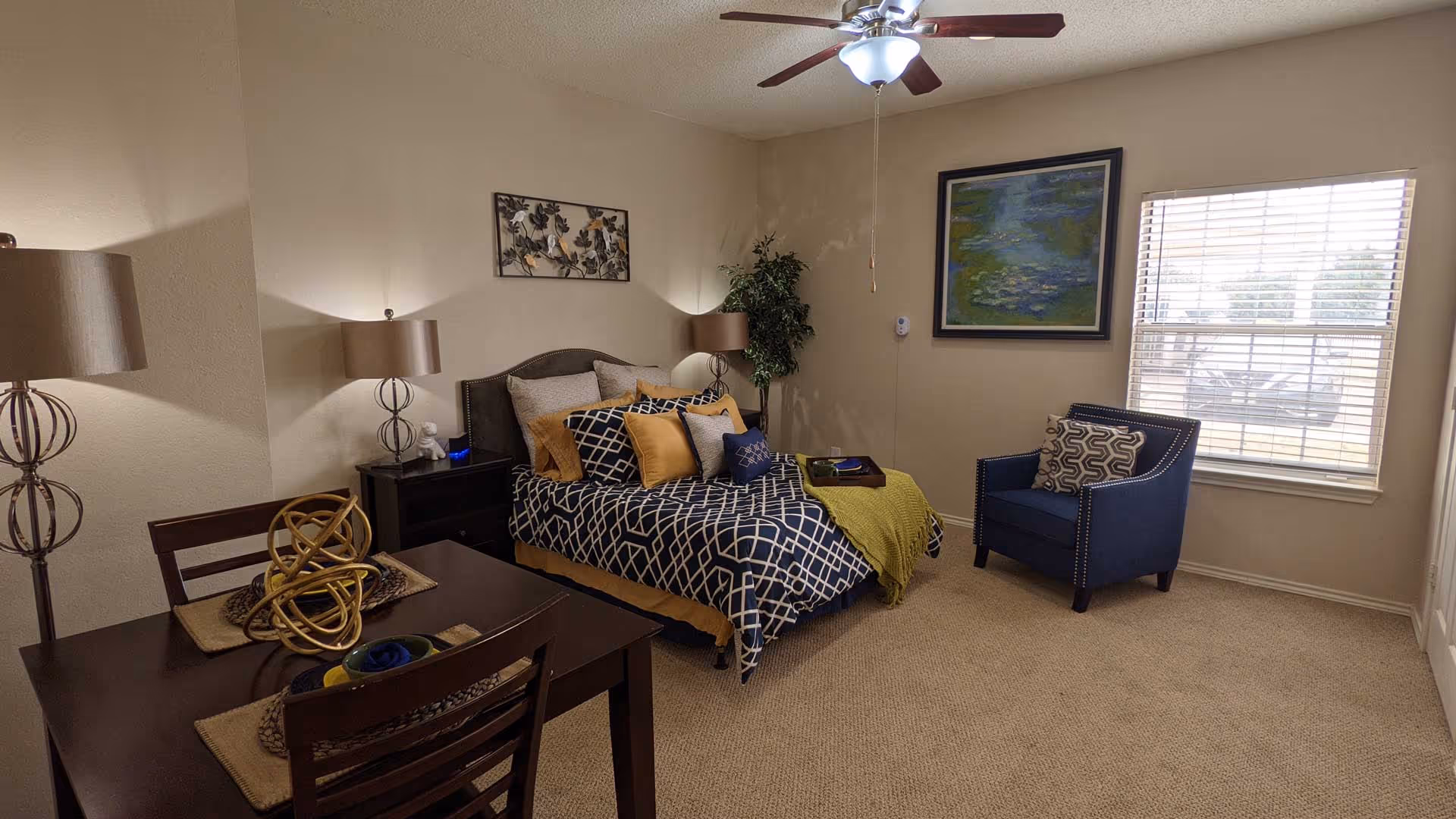 A cozy bedroom with a bed dressed in navy blue and white geometric patterned bedding and multiple pillows in shades of yellow and gray. There are two bedside tables with matching lamps, a small dining table with two chairs, a blue armchair with a patterned cushion near a window with blinds, a ceiling fan with lights, and wall art above the bed and near the window. The room has beige walls and carpeted flooring.