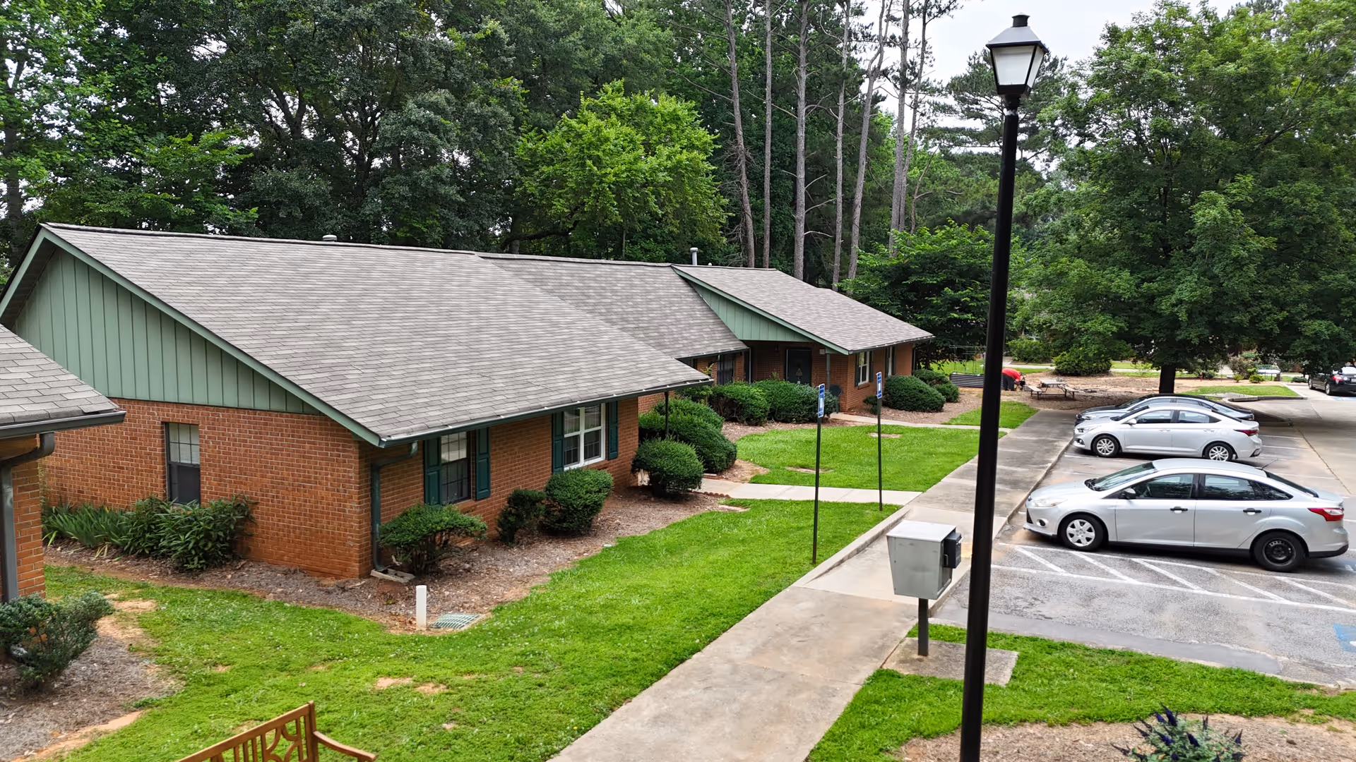 Exterior view of single-story brick residential buildings with green trim and gray shingled roofs, surrounded by well-maintained grass, bushes, and trees. A paved walkway and parking lot with several parked cars are visible, along with a tall black street lamp.