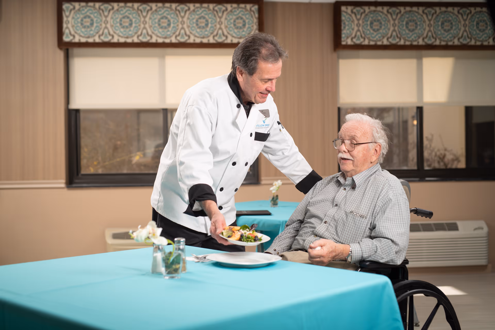 A chef in a white uniform is serving a plate of food to an elderly man in a wheelchair seated at a table with a turquoise tablecloth in a dining area with windows and patterned valances.