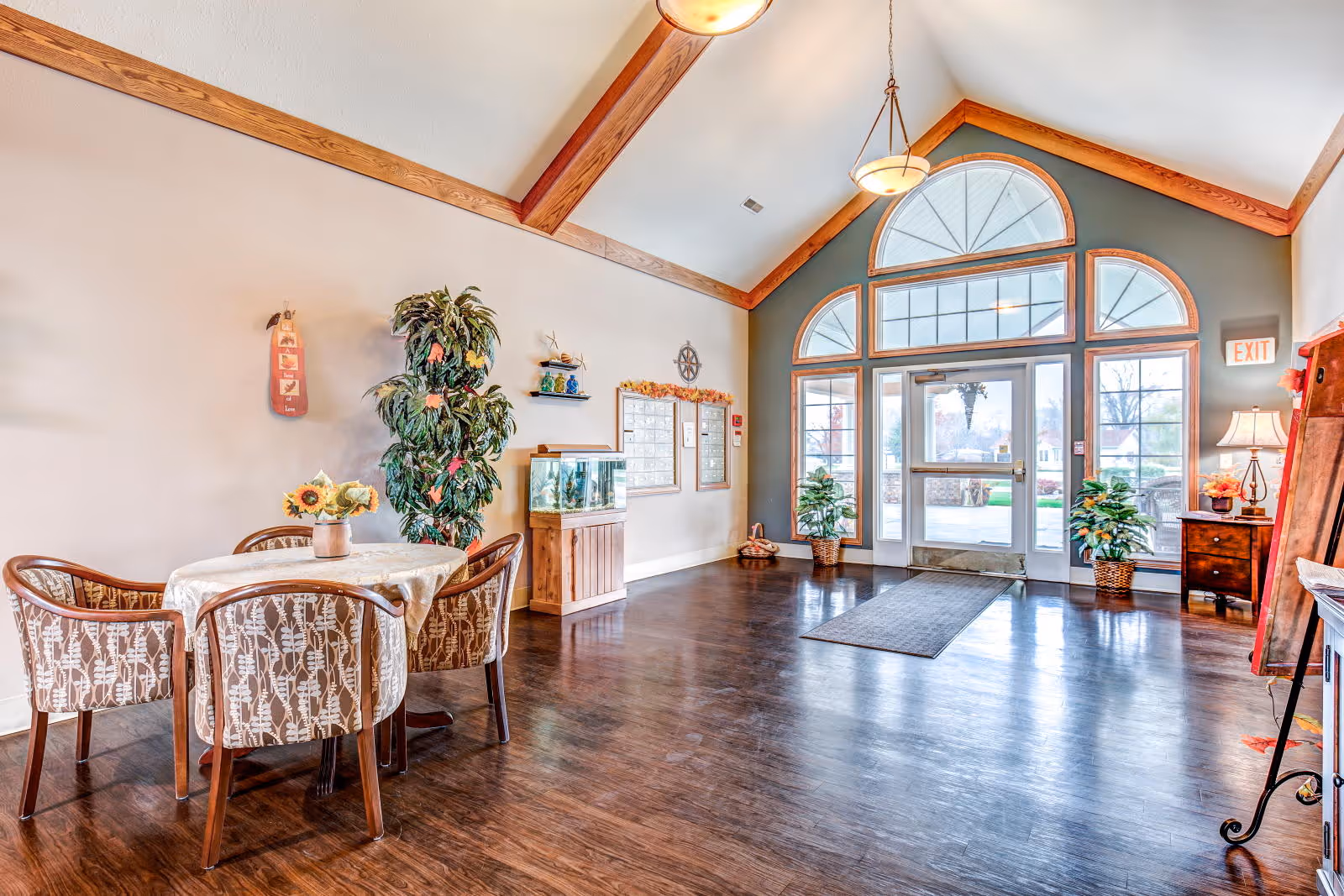 A bright and spacious interior common area with high vaulted ceilings and wooden beams. There is a round table with four patterned chairs and a vase of sunflowers on top. A large potted plant and a fish tank on a wooden stand are against the wall. The far wall features large arched windows and a glass door leading outside. The floor is dark wood, and there are two hanging light fixtures from the ceiling. Additional plants and a small table with a lamp are near the entrance.