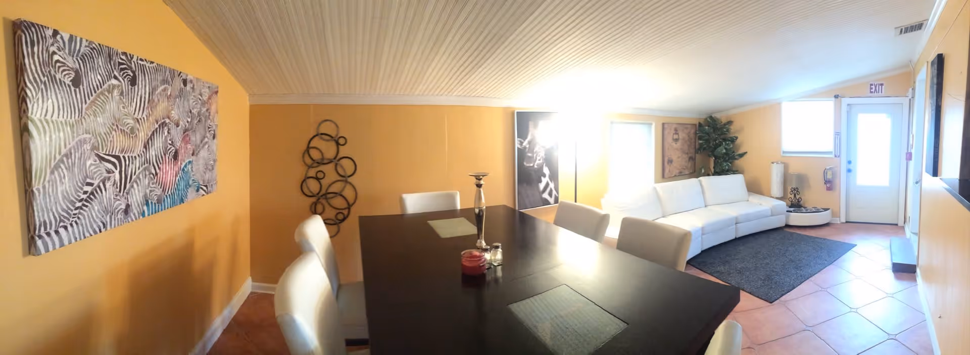 A bright room with yellow walls featuring a dark wooden dining table surrounded by white chairs. On the left wall, there is a large painting of zebras. The room also has a white sectional sofa near a door with an exit sign above it. There is a decorative metal wall art piece and a tall plant in the corner, with a dark rug on the tiled floor.