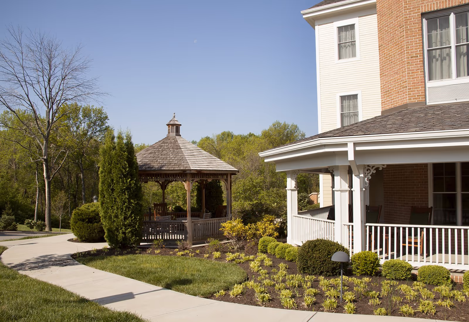 Landscaped outdoor area featuring a wooden gazebo, a covered porch on a multi-story building, and a winding concrete pathway.