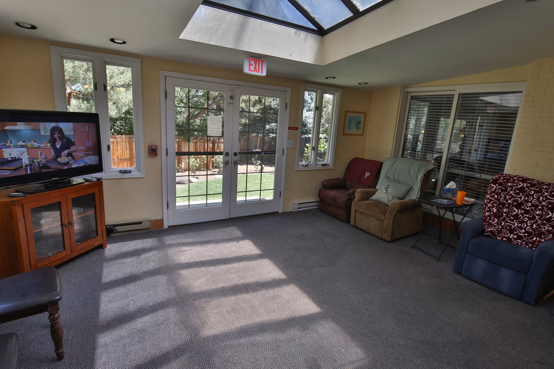 A cozy living room area with three armchairs, a TV on a wooden stand showing a cooking show, large windows and glass doors leading to a garden outside, and a skylight letting in natural light.
