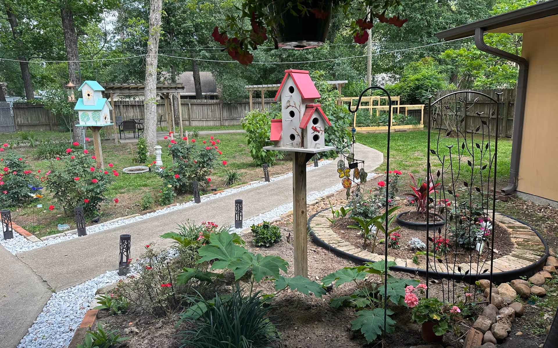 Colorful garden courtyard with birdhouses, pathways, and flowering plants in a fenced yard.