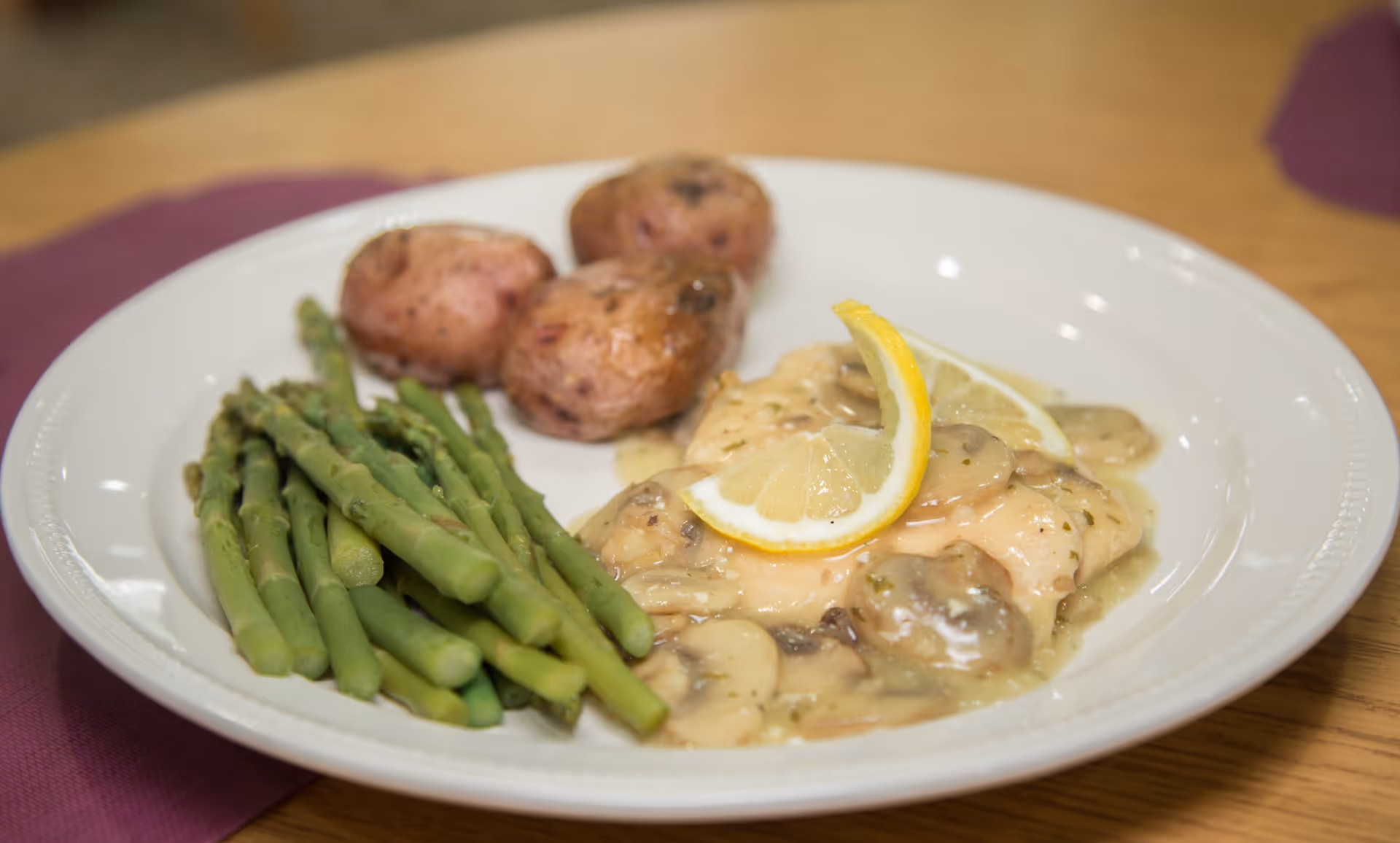 A white plate with cooked asparagus, three small roasted potatoes, and a piece of chicken covered in mushroom sauce, garnished with two lemon slices, placed on a wooden table with a purple napkin underneath.