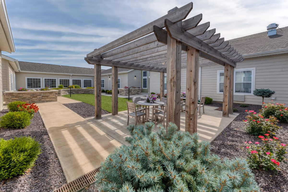 Outdoor courtyard area at High Point Residence Quincy featuring a wooden pergola with a round table and chairs underneath. The courtyard is surrounded by beige buildings with windows, landscaped with green shrubs, flowering plants, and a concrete walkway.