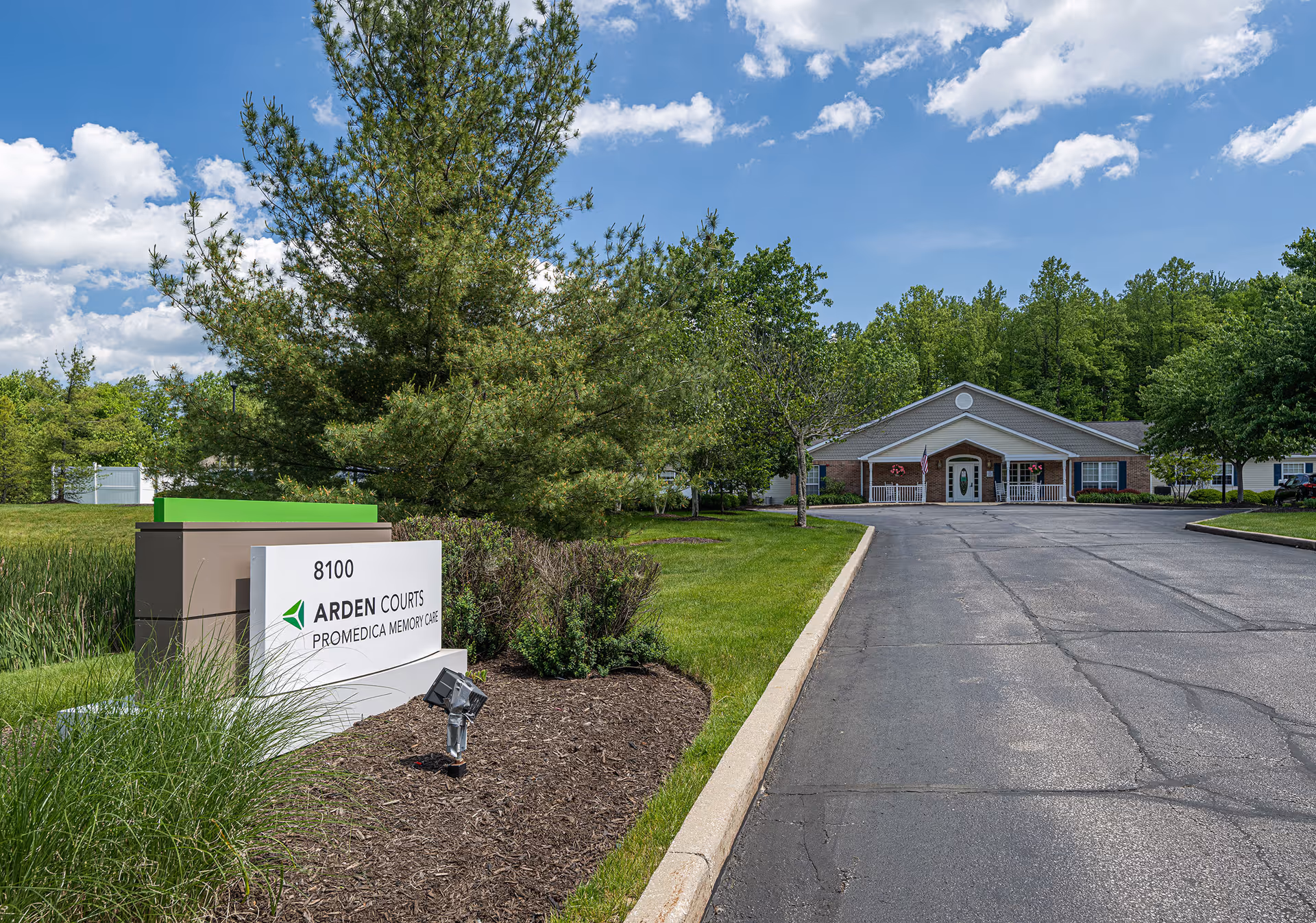 Exterior view of Arden Courts - ProMedica Memory Care Community in Chagrin Falls, showing the driveway leading to the building entrance surrounded by green trees and grass under a partly cloudy sky. A sign with the facility name and address is visible in the foreground.