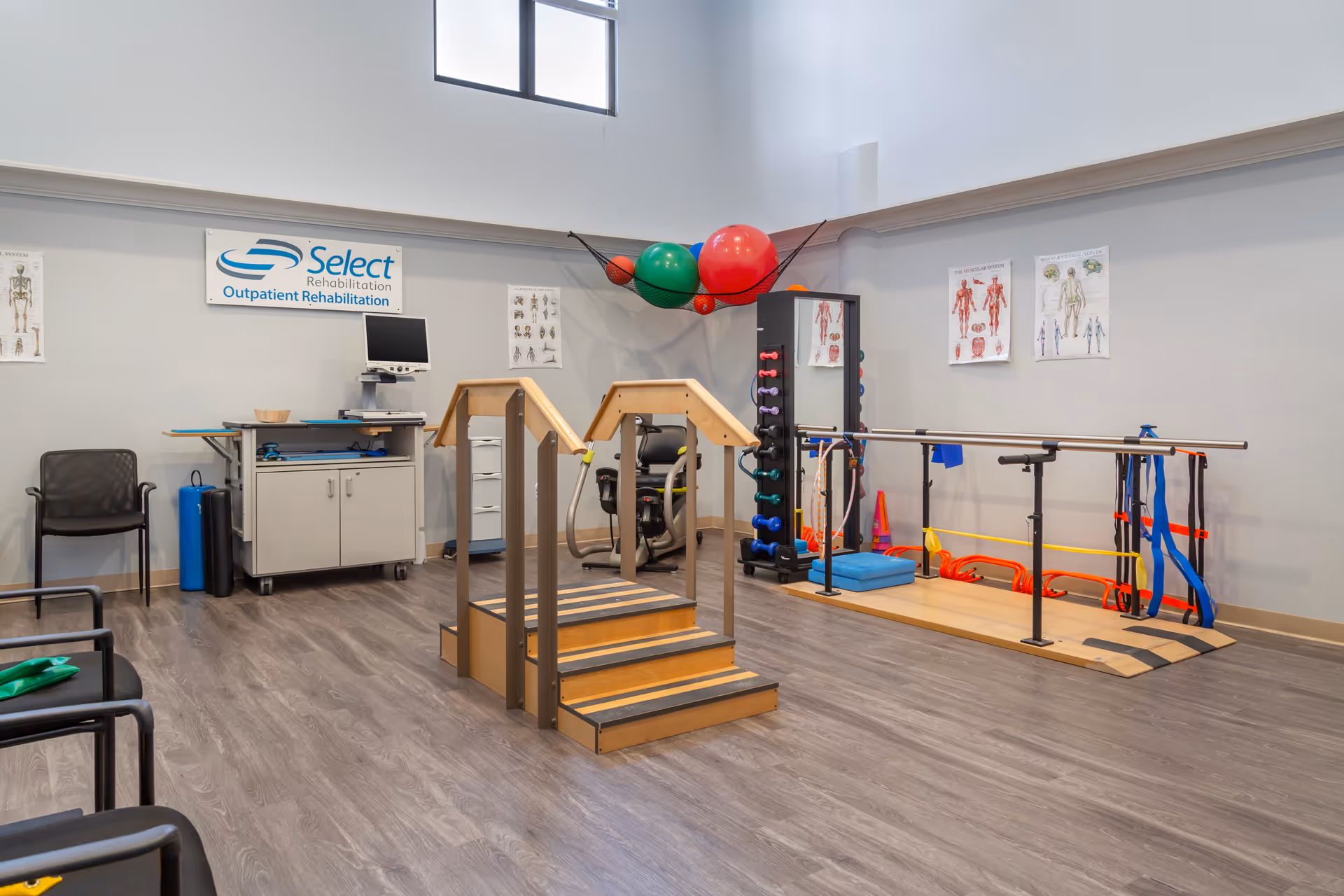 A rehabilitation room with exercise equipment including a set of wooden stairs with handrails, parallel bars with resistance bands, a rack of colorful dumbbells, and large exercise balls stored in a net hanging from the wall. There are anatomical posters on the walls and a workstation with a computer monitor. Chairs are arranged along the left side of the room.