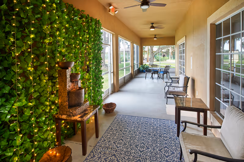 A screened-in covered porch with chairs and tables, a decorative ivy wall strung with lights, and a small tabletop fountain.