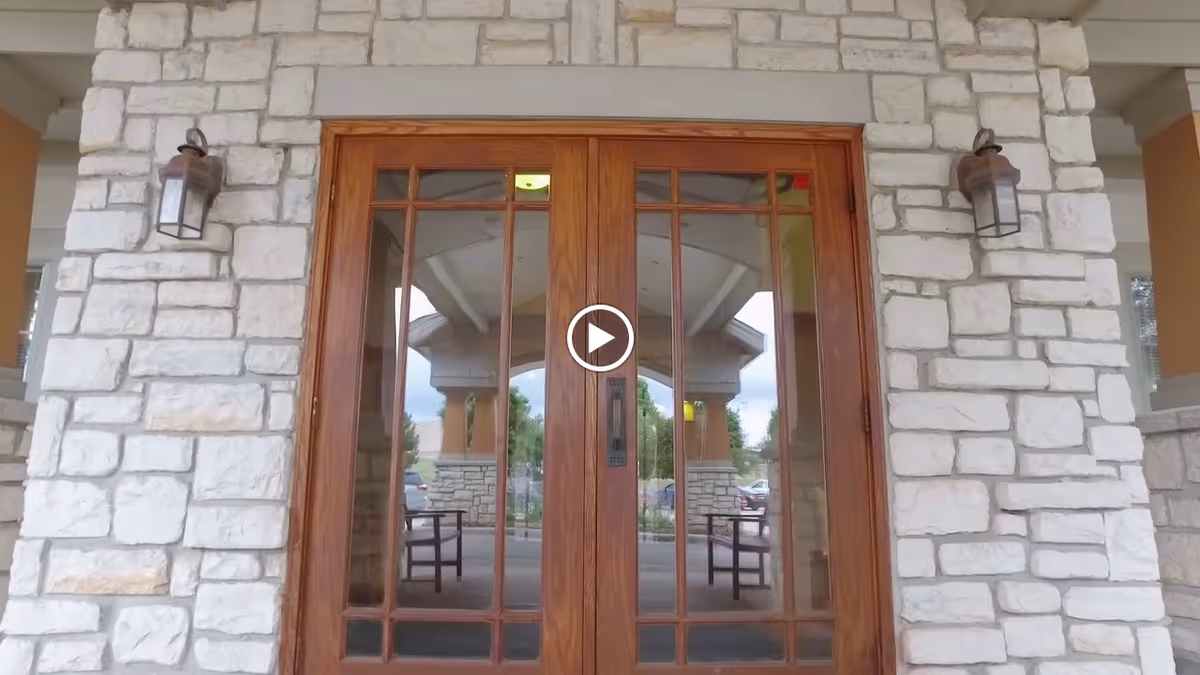Double wooden doors with glass panels set in a stone wall, with two outdoor wall lanterns on either side. The glass reflects an outdoor covered entrance area with stone pillars and some parked cars in the background.