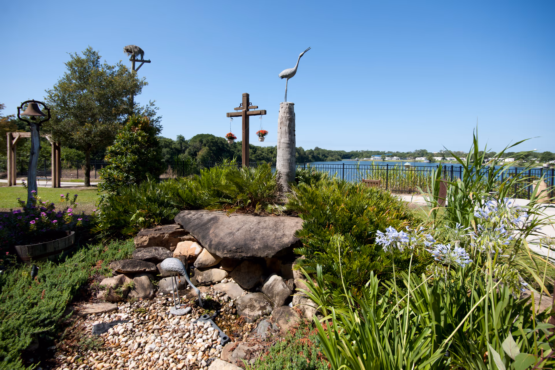 A landscaped outdoor garden area with various green plants, flowers, and decorative bird sculptures. In the background, there is a body of water with a clear blue sky above. The garden includes rocks and a small gravel area, with a wooden cross structure and hanging flower pots visible. Trees and a fence line the perimeter.