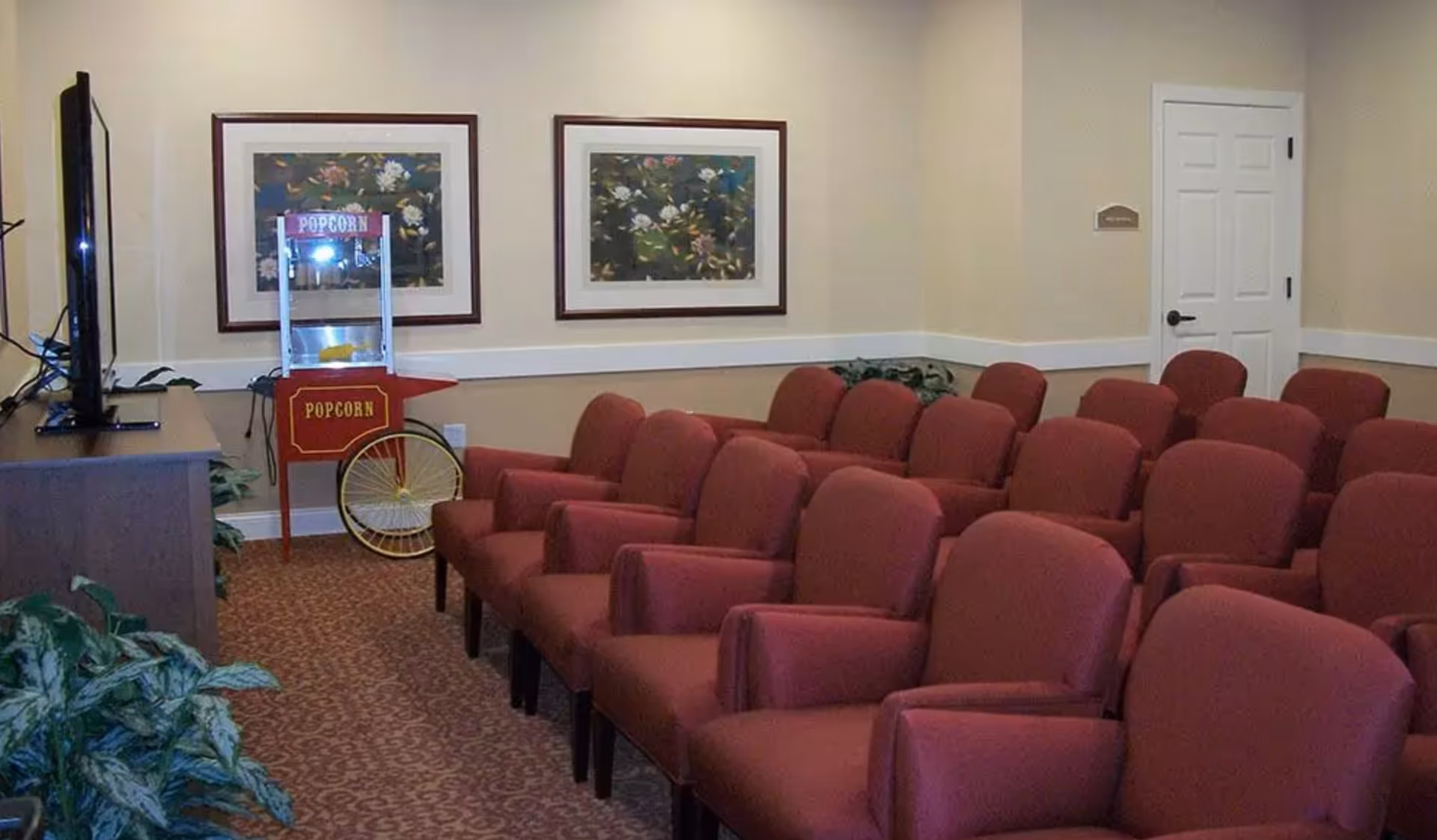 A small theater room with rows of red upholstered chairs facing a television screen. There is a vintage-style popcorn machine on wheels near the front, and two framed floral paintings hang on the beige walls. A white door is visible in the background.