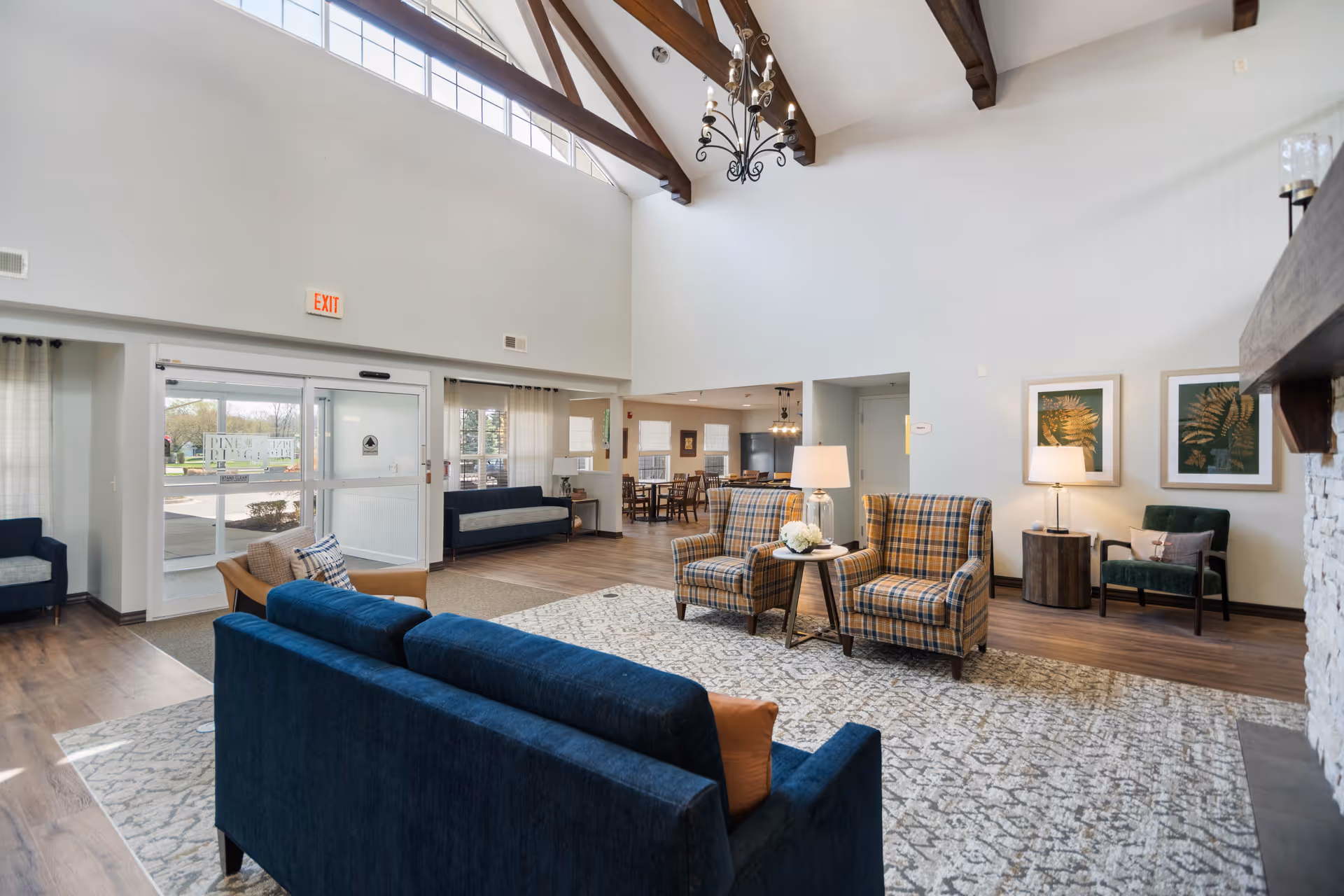 A spacious and well-lit living room area with high ceilings and exposed wooden beams. The room features a blue sofa, two plaid armchairs with a small round table between them, a green armchair, and a large patterned rug. There are two framed botanical prints on the wall, a stone fireplace on the right, and large windows letting in natural light. In the background, there is a dining area with tables and chairs, and glass doors leading outside with a sign visible through the glass.