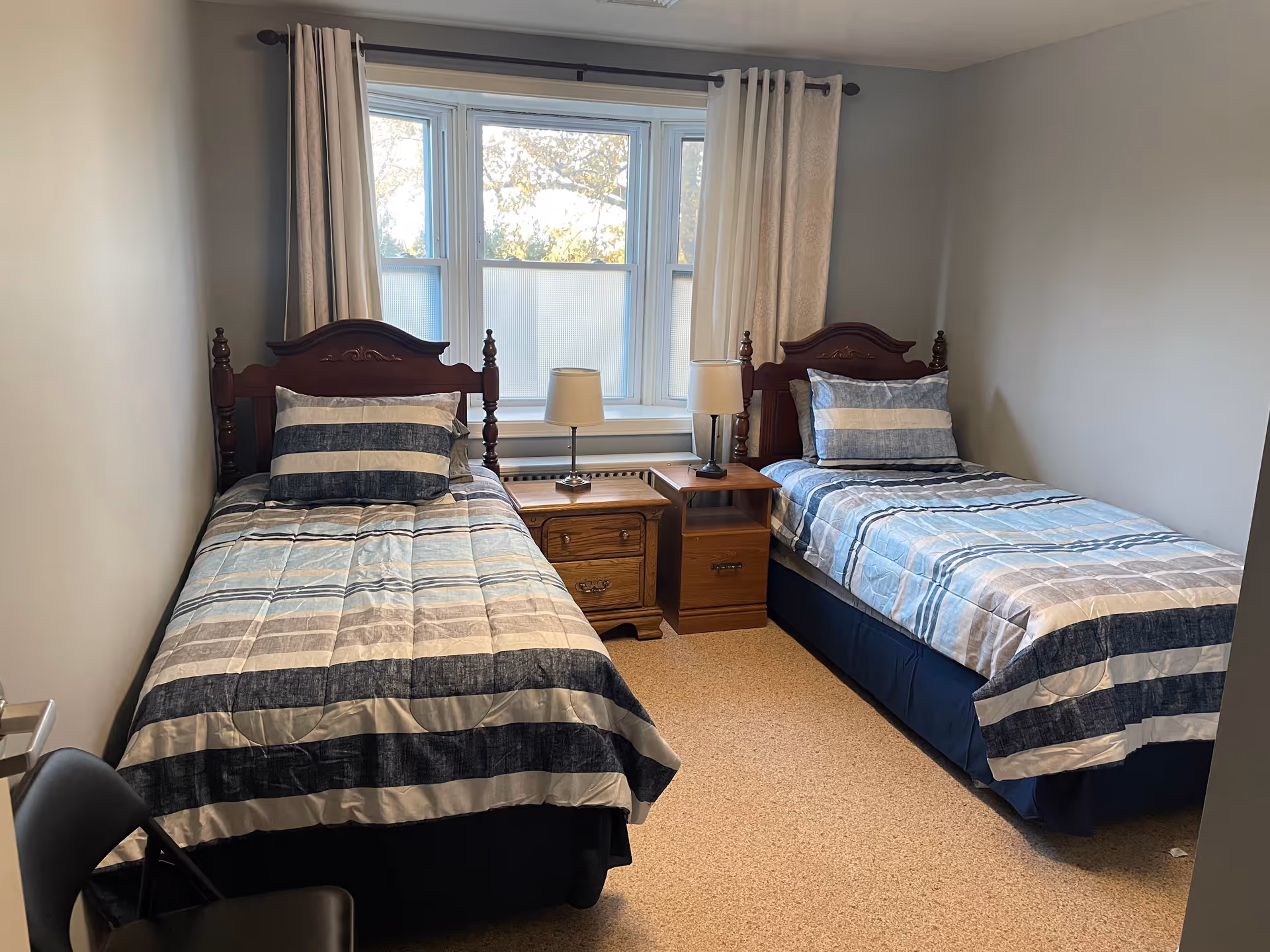 Bedroom with two twin beds featuring striped bedding, wooden headboards, a central nightstand with a lamp, and a window with curtains.