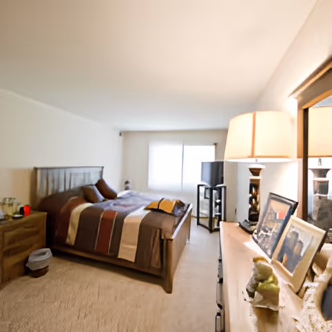 A cozy bedroom with a wooden bed frame and a striped bedspread in shades of brown, beige, and red. There is a wooden nightstand with items on it to the left of the bed, and a dresser with a lamp, framed photos, and decorative items on the right. A window with closed blinds is visible at the far end of the room, and a TV is placed on a stand near the window.