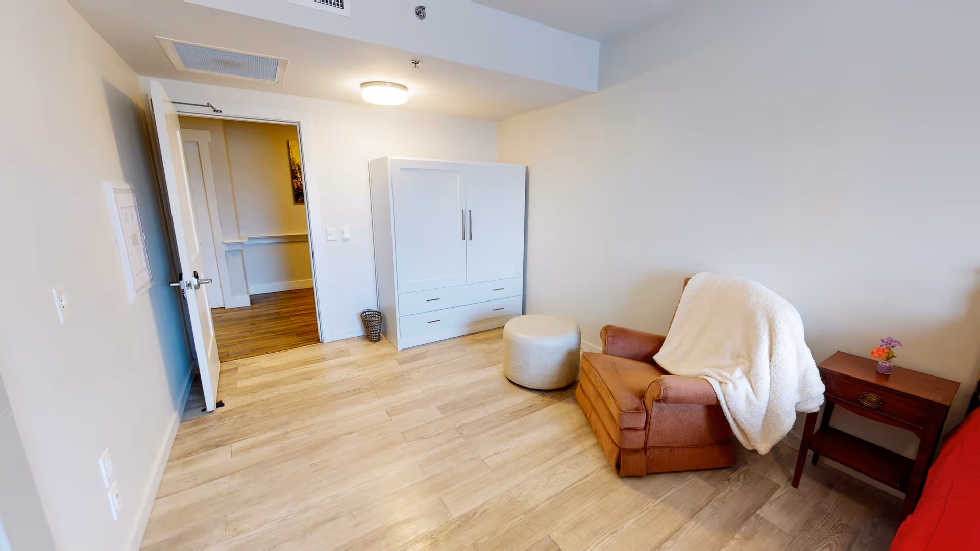 A small room with light wood flooring and white walls. There is a white cabinet with drawers against the far wall, a round beige ottoman in front of it, and a brown armchair with a white blanket draped over it to the right. Next to the armchair is a small wooden side table with a small potted plant. An open door leads to another room with wood flooring and a piece of wall art visible.