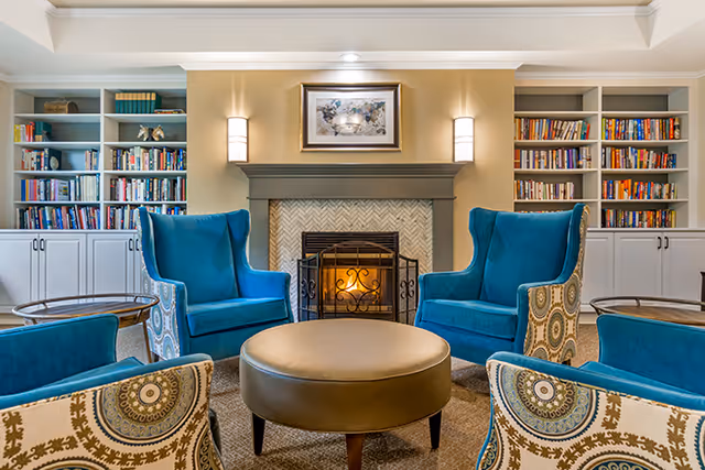 A cozy living room area with a lit fireplace in the center, flanked by built-in bookshelves filled with books. Four blue upholstered armchairs with patterned backs surround a round brown ottoman coffee table. The walls are painted beige, and there is a framed picture hanging above the fireplace with two wall sconces on either side.