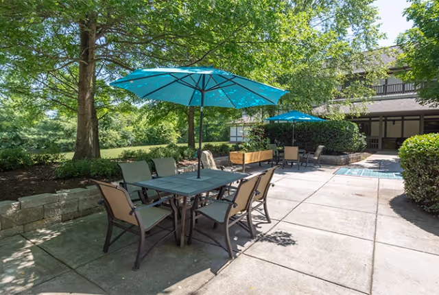 Sunlit outdoor patio with tables, chairs, and teal umbrellas adjacent to a landscaped lawn and building.