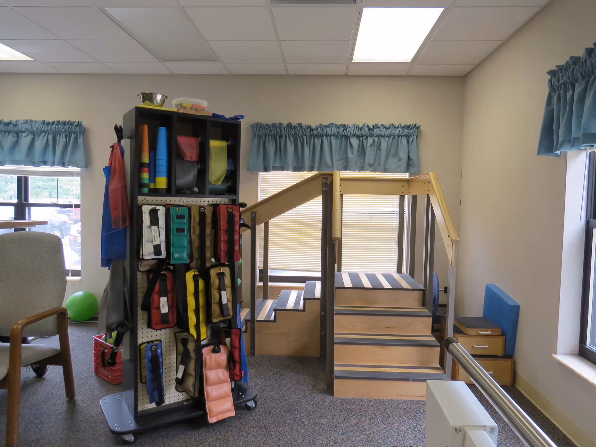 Interior therapy room with wooden exercise stairs, a rolling equipment rack stocked with weighted cuffs and therapy supplies, a chair, and windows with blue valances.