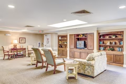 A bright and spacious living room area in a senior living facility featuring a floral patterned sofa, two wooden armchairs with cushions, a small round side table, built-in wooden shelves with books and decorative items, and a television set in a wooden entertainment center. The room has beige walls, carpeted floor, recessed ceiling lights, and a table with two chairs and a lamp in the background.