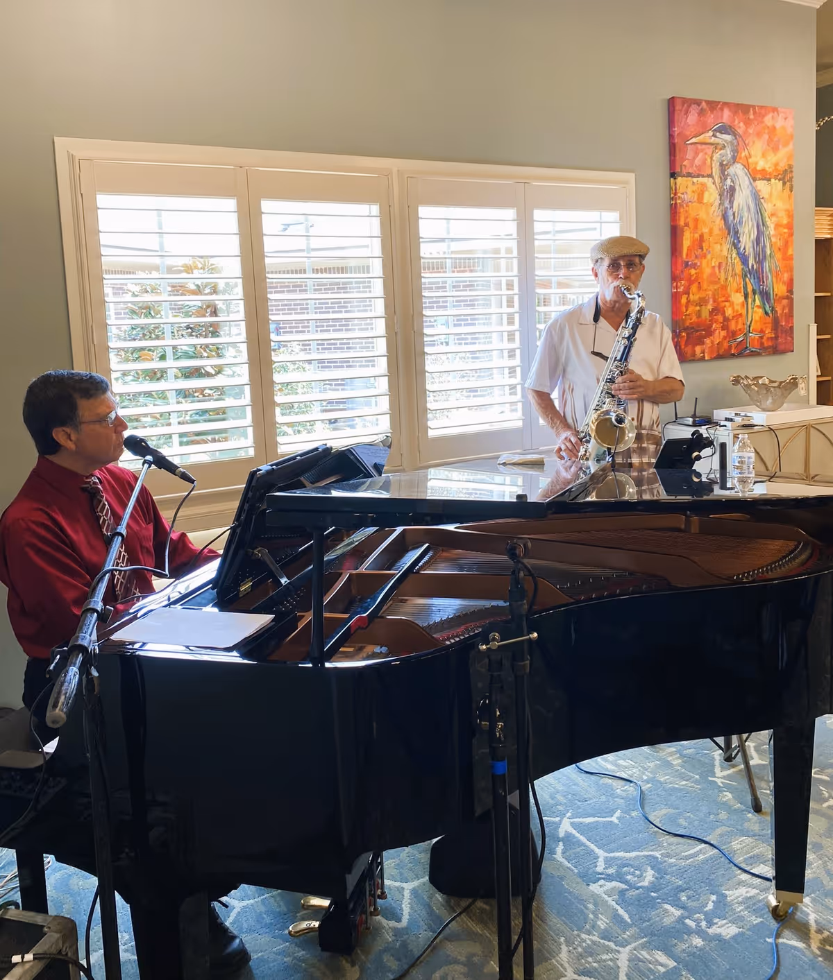 Two men performing music indoors; one man in a red shirt and tie is playing a grand piano and singing into a microphone, while the other man wearing a hat and white shirt is playing a saxophone. They are in a room with large windows with white shutters and a colorful painting of a bird on the wall.