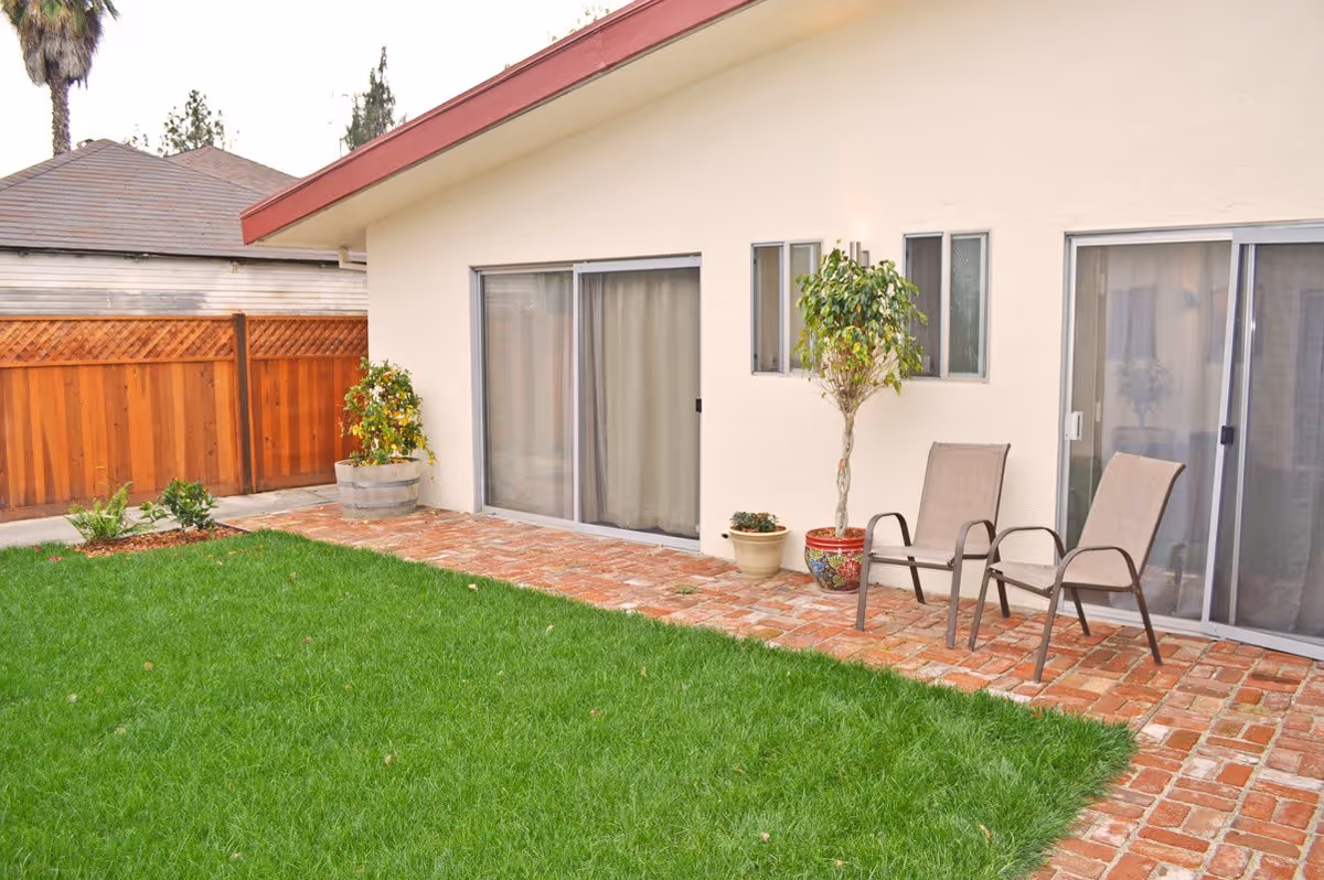 A backyard area with a green lawn, brick patio, two beige outdoor chairs, and potted plants against the exterior wall of a light-colored building with sliding glass doors and small windows.