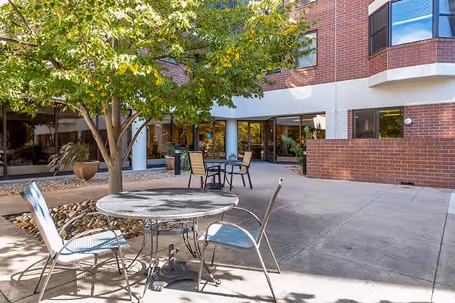 Outdoor patio area at Brookdale Meridian Englewood with metal tables and chairs, a tree providing shade, and a brick building with large windows in the background.