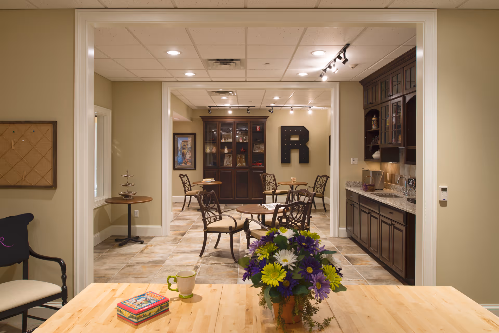 Interior view of a senior living facility common area with a wooden table in the foreground holding a colorful flower arrangement, a coffee mug, and a decorative tin box. Beyond the table, there are several small round tables with chairs, a dark wooden cabinet with glass doors, and a large letter 'R' mounted on the wall. The room has beige walls, tiled floors, and recessed lighting.