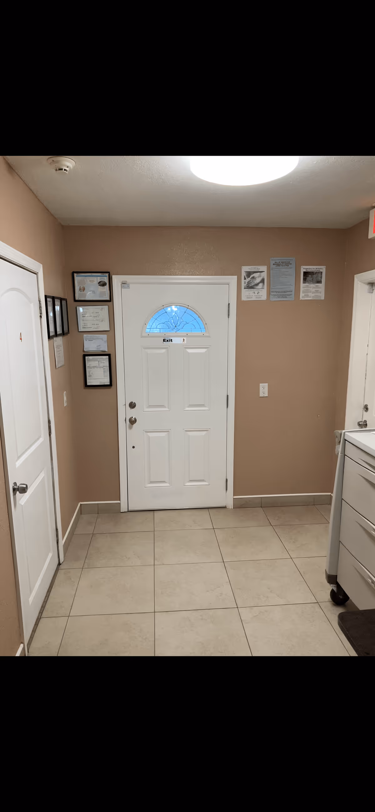 Interior view of a hallway in a facility with beige walls and tiled floor. A white door with a small decorative window and an 'Exit' sign is centered on the far wall. To the left, there is another white door labeled with the number 4 and several framed certificates or notices on the wall. On the right side, part of a white cabinet or cart with drawers is visible. The ceiling has a round light fixture and a smoke detector.