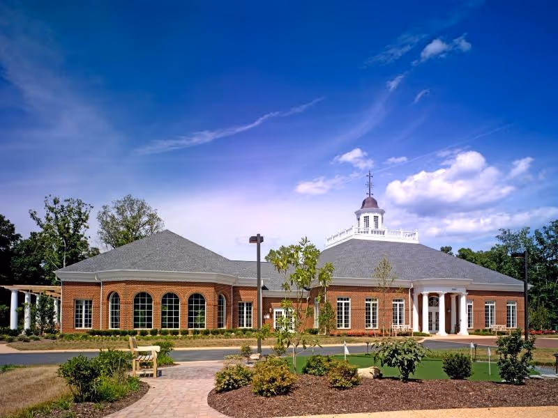 Brick single-story senior living building with a columned entrance, a cupola on the roof, and landscaped grounds under a blue sky.