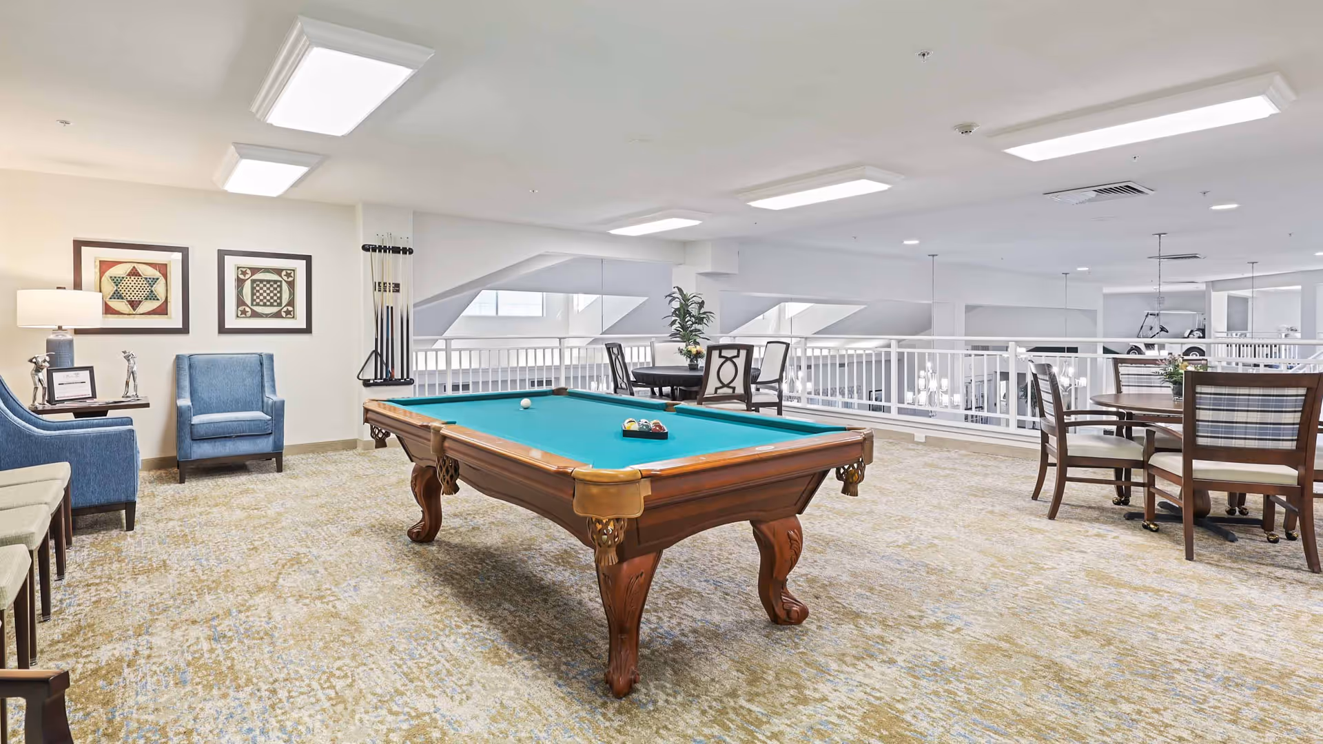 A bright and spacious recreational room featuring a wooden pool table with billiard balls arranged on the green felt surface. The room has beige patterned carpet flooring and white walls. There are two blue upholstered armchairs with a small side table and lamp between them on the left side. On the right side, there are two round tables with chairs around them. The room overlooks a lower level through white railings, and the ceiling has multiple rectangular recessed lights.