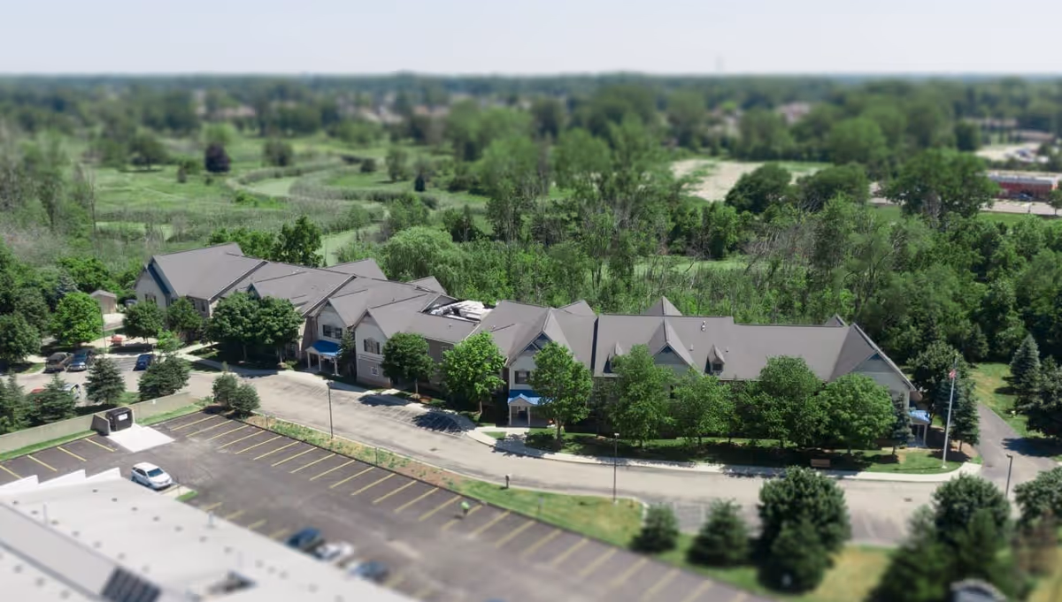 Aerial view of a large senior living facility building surrounded by trees and greenery, with a parking lot in the foreground and a sprawling landscape in the background.