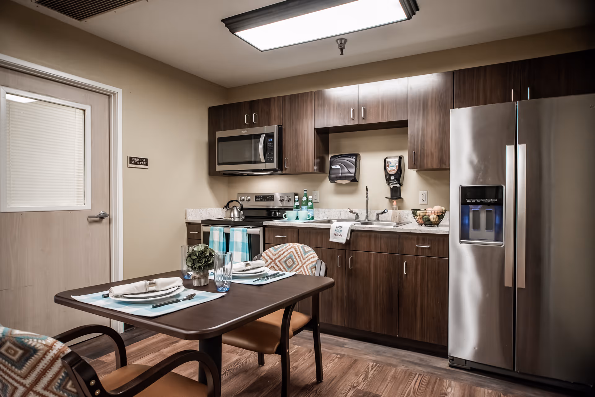Interior view of a kitchen area in a nursing and rehabilitation center featuring dark wood cabinets, a stainless steel refrigerator, microwave, stove, and sink. A small dining table with two chairs is set with placemats, napkins, and glassware. The room has wood flooring and a door labeled 'Director of Therapy'.