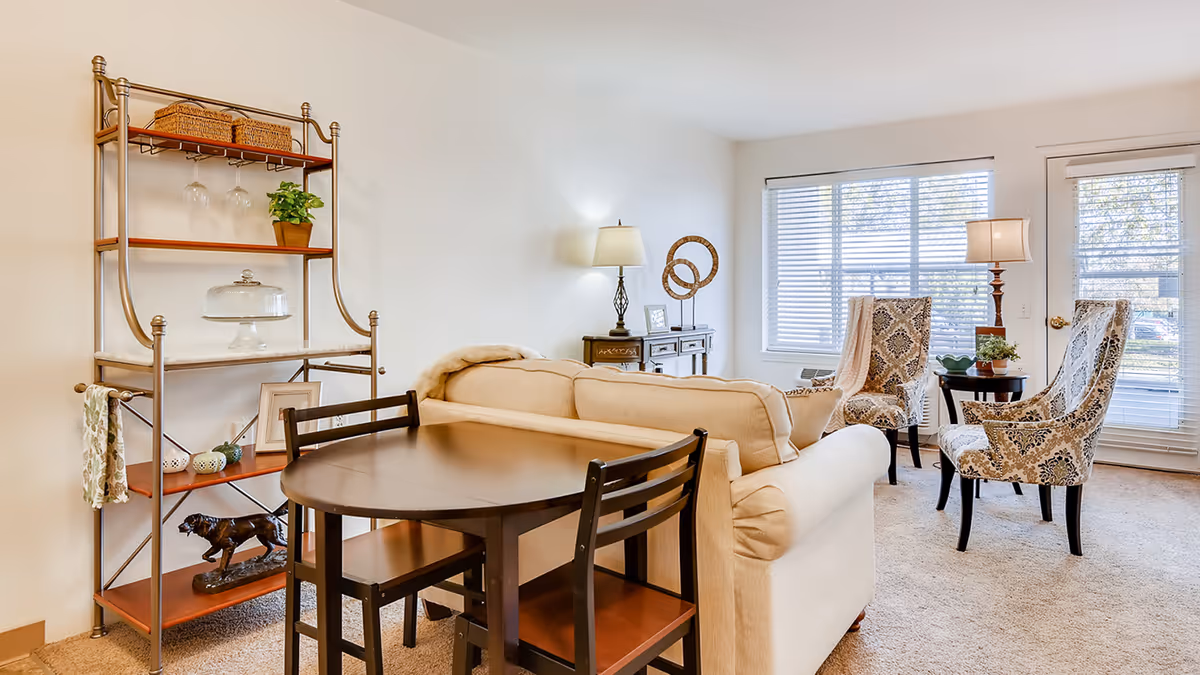 A bright living room with a beige sofa, two patterned armchairs, a small round table with plants, a wooden dining table with chairs, a metal shelving unit with decorative items, and a window with blinds letting in natural light.