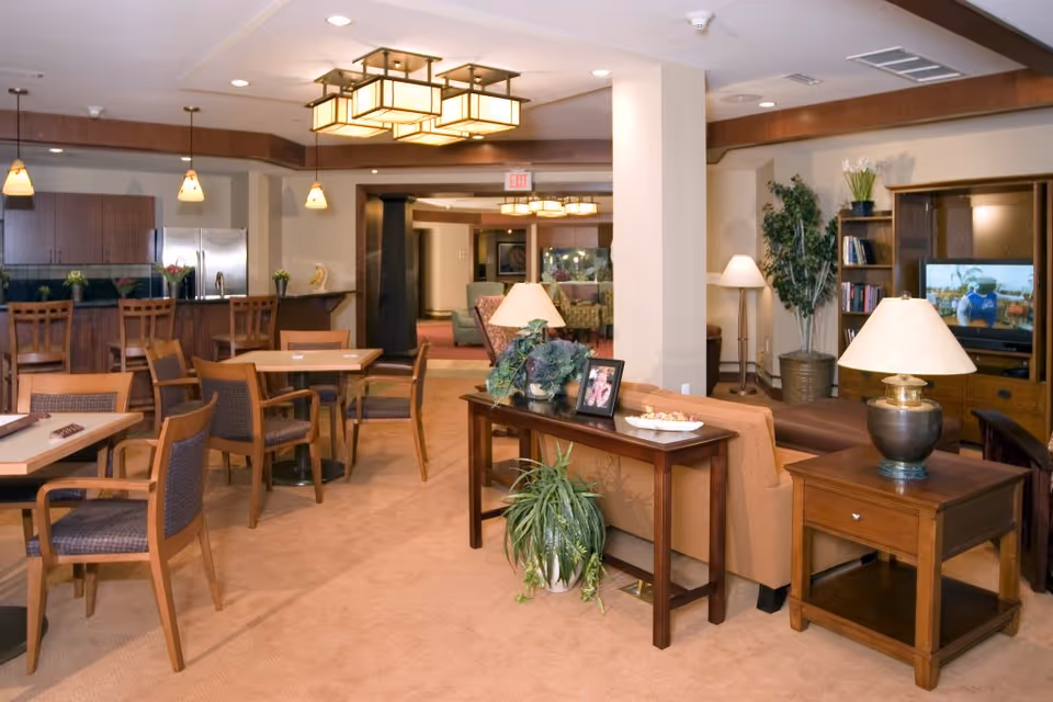 A spacious and well-lit common area in a senior living facility featuring a seating area with a sofa, armchairs, and wooden tables with lamps. In the background, there is a kitchen area with a refrigerator, cabinets, and a counter with bar stools. The room is decorated with plants, framed photos, and has a television on a wooden entertainment center.