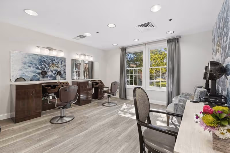 A bright and modern hair salon area inside The Forum At Knightsbridge facility featuring two styling stations with mirrors, chairs, and hair dryers. There is a large window with gray curtains letting in natural light, and a small seating area with chairs and a table with flowers.