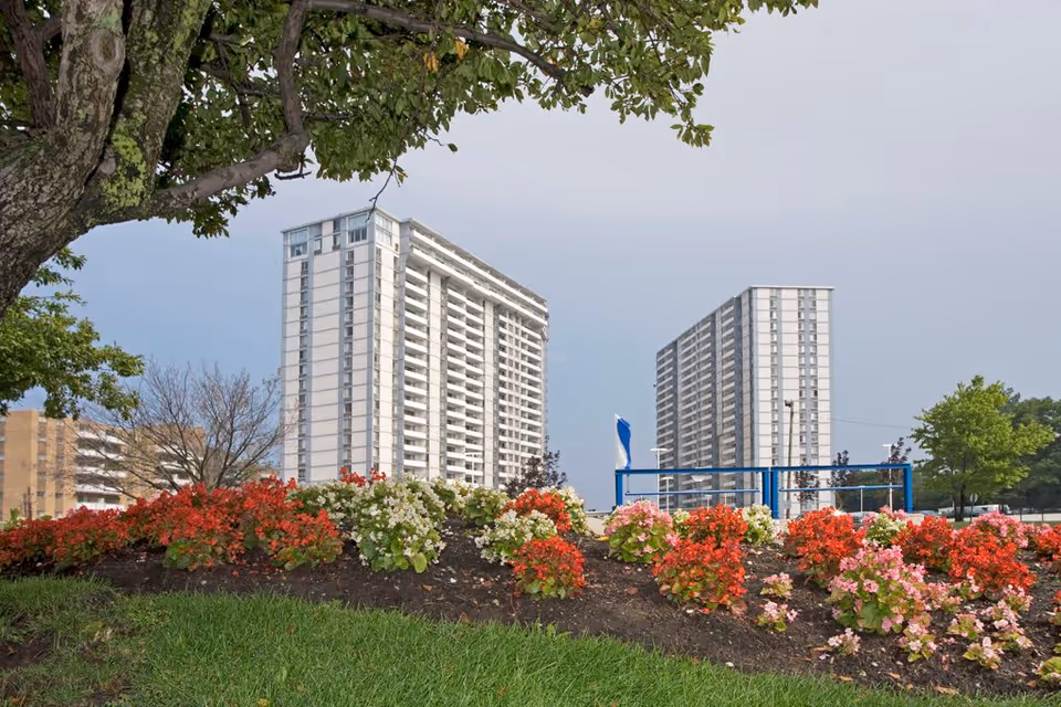 Two high-rise residential buildings framed by a tree and colorful flowerbeds in the foreground.