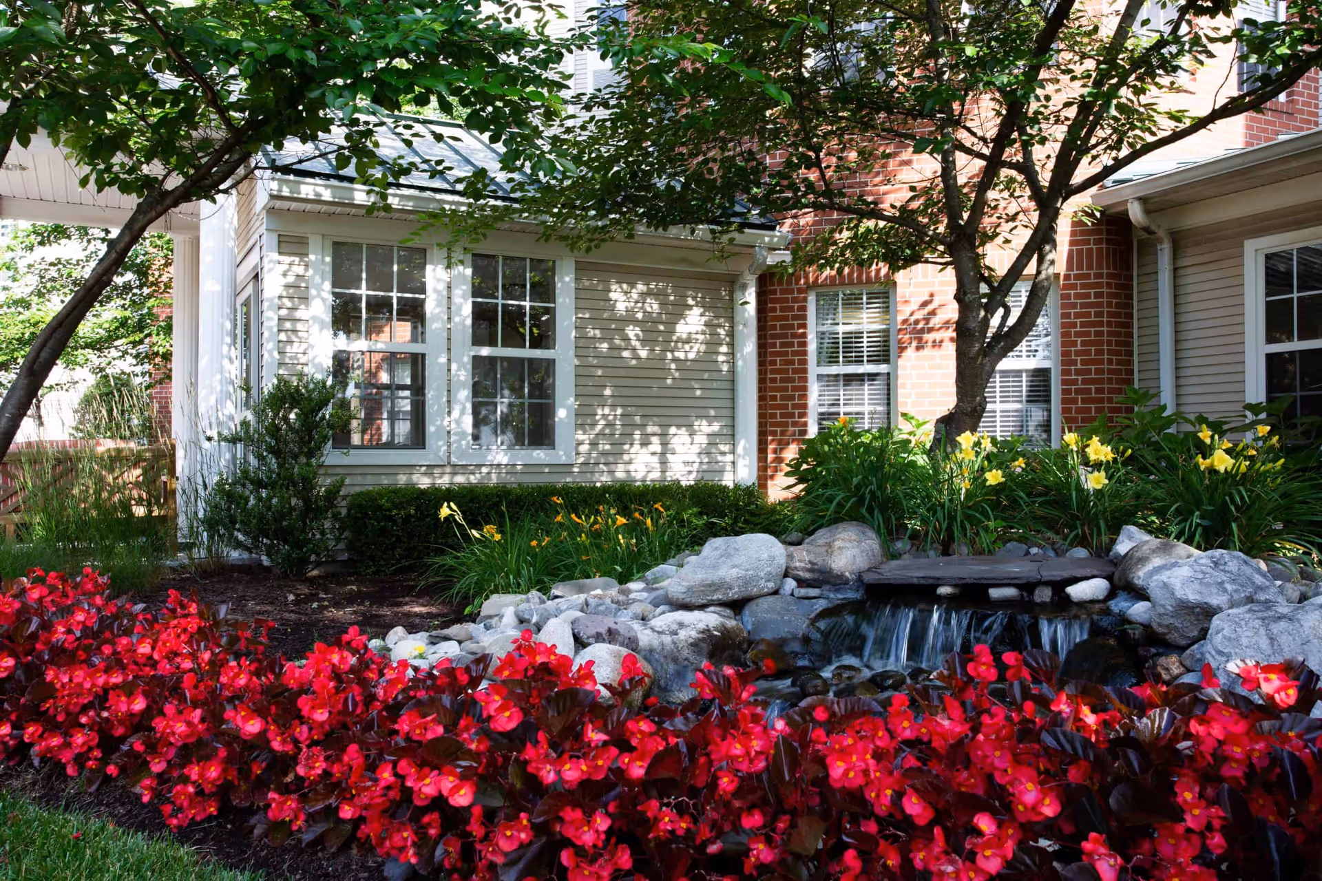 Exterior garden with red flowers, a small rock waterfall pond, trees, and building windows.