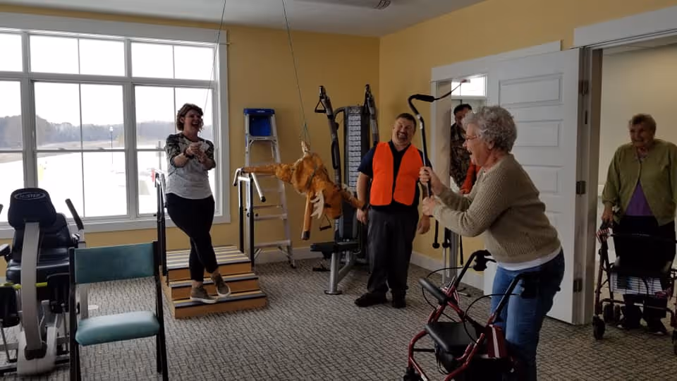 A group of elderly people and a younger woman are in a fitness room with exercise equipment. One elderly woman with a walker is swinging a stick at a hanging piñata shaped like a moose, while others watch and laugh. The room has large windows and yellow walls.