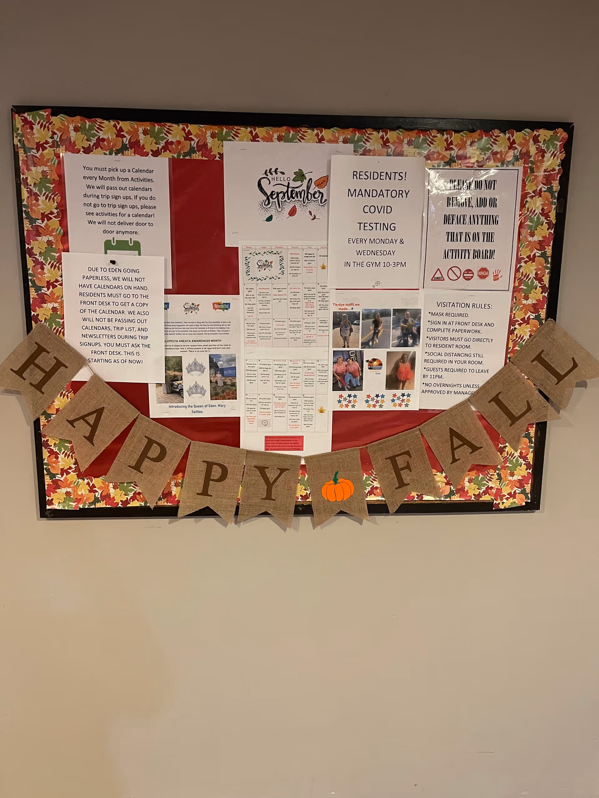 A bulletin board decorated with a fall-themed border and a burlap banner that reads 'HAPPY FALL' with a pumpkin image. The board contains various notices including a calendar pickup reminder, COVID testing schedule, visitation rules, and a sign requesting not to remove or deface anything on the board. The notices are printed on white paper with some colorful graphics and photos.