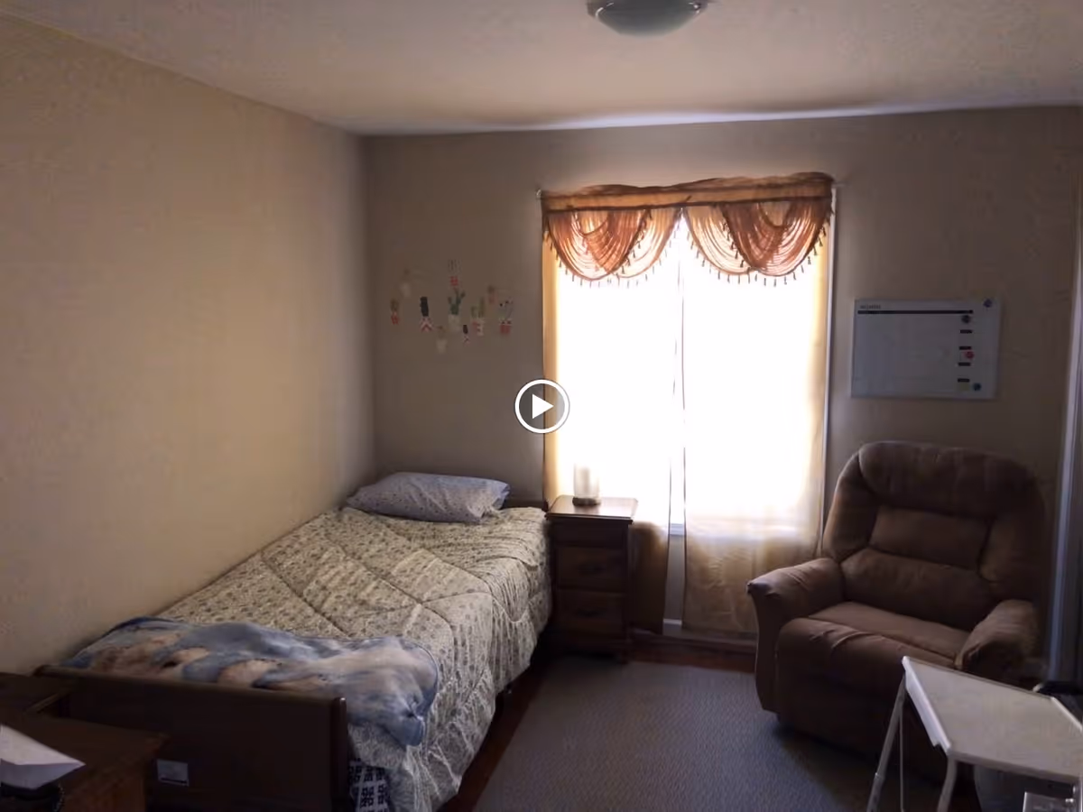 A small bedroom with a single bed covered with a patterned quilt and a pillow. Next to the bed is a wooden nightstand with a candle on it. A window with sheer curtains and a decorative valance lets in light. There is a comfortable recliner chair and a whiteboard on the wall. The room has beige walls and carpeted floor.